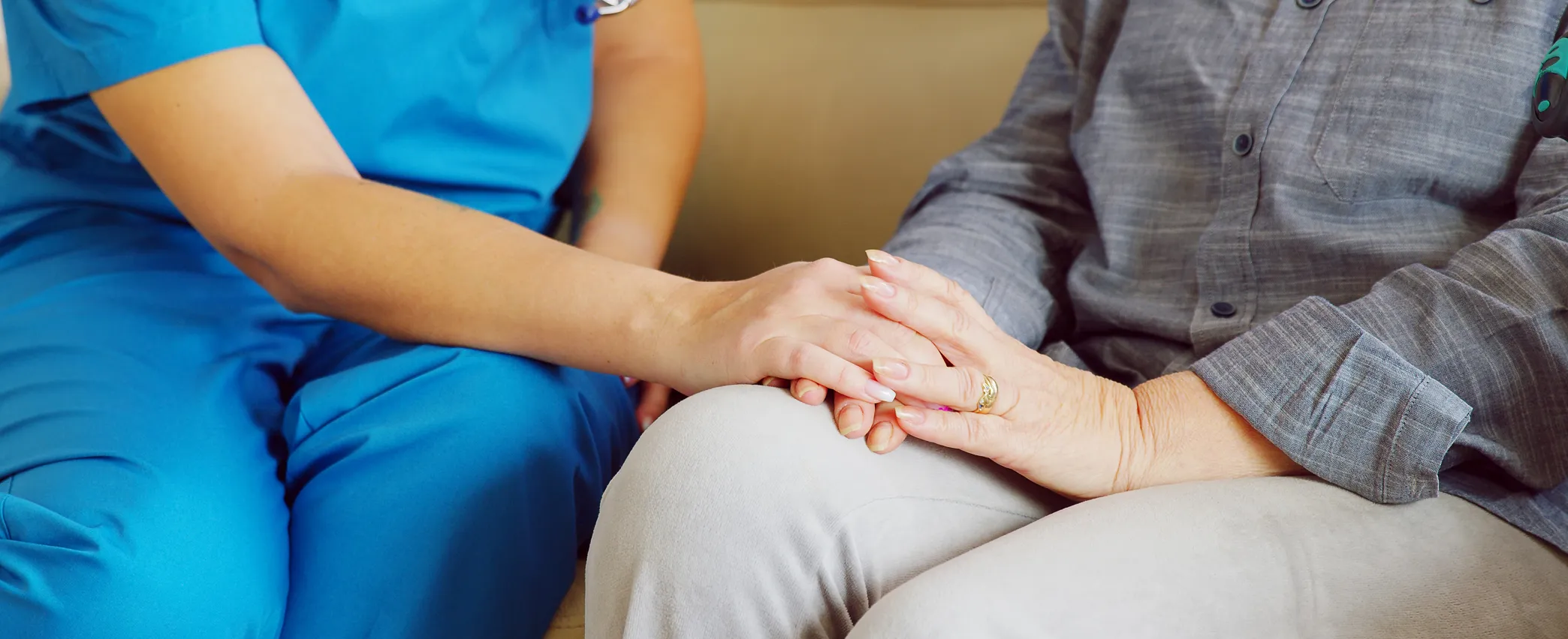 Healthcare professional in blue scrubs holding the hand of an elderly patient wearing a gray shirt and light pants.