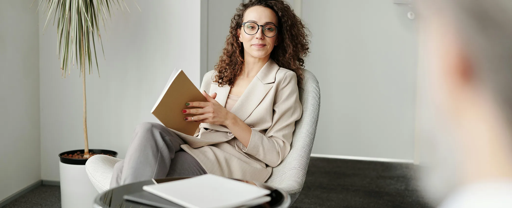 A woman with curly hair and glasses sits in a chair holding a notebook, with a potted plant in the background.