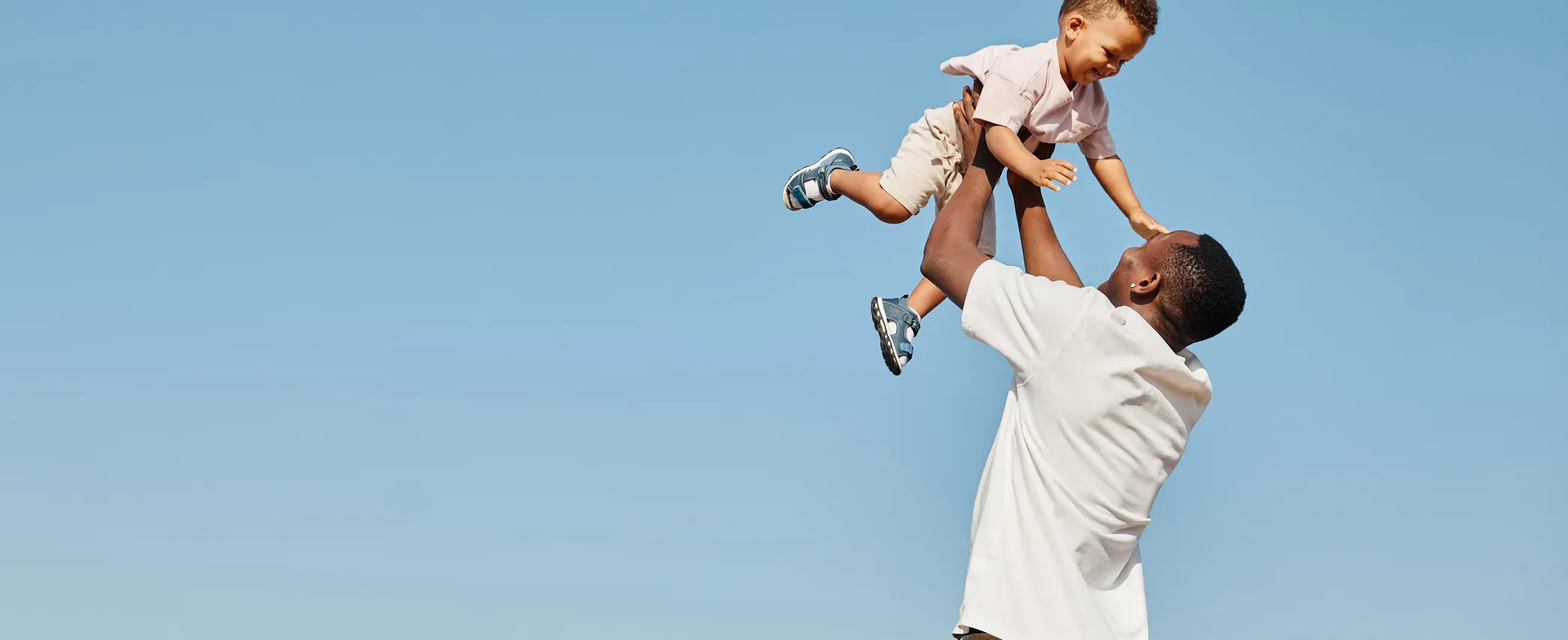 Man lifting a smiling child in the air against a clear blue sky.