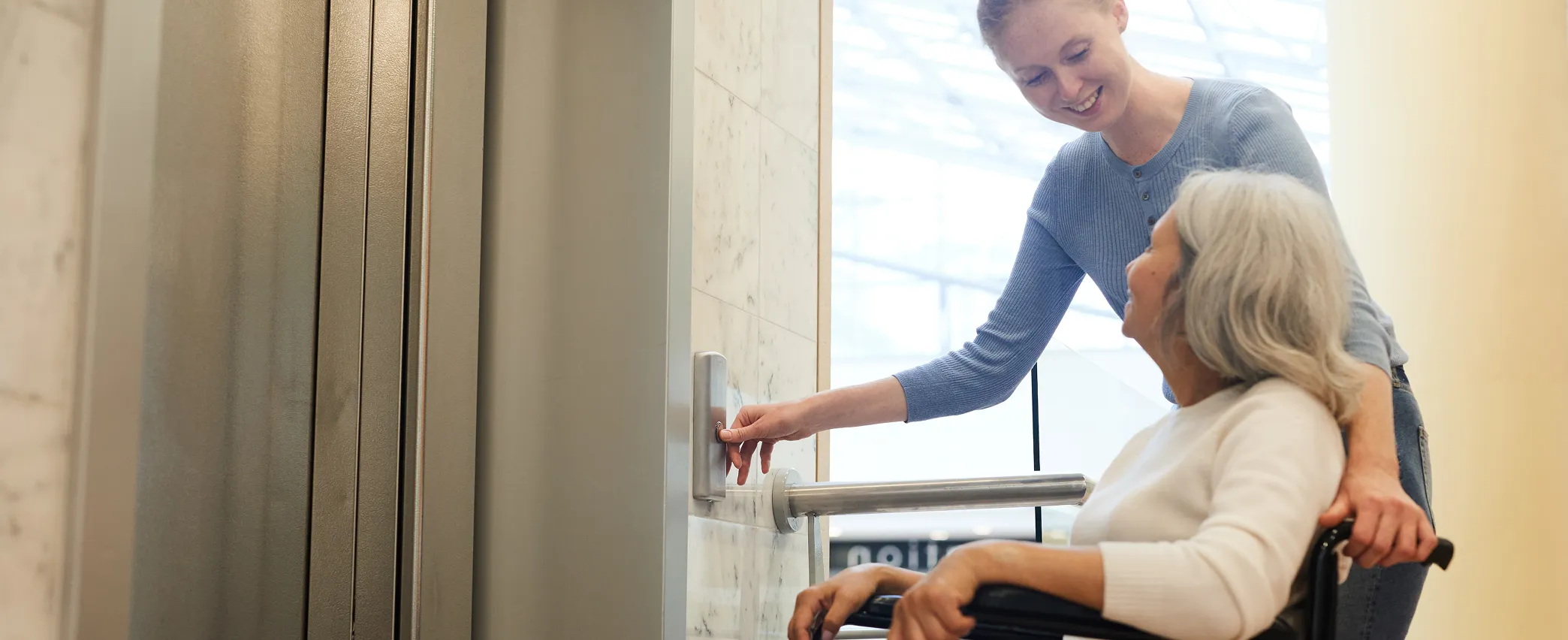 Young woman smiling while pressing an elevator button as she assists an older woman in a wheelchair.