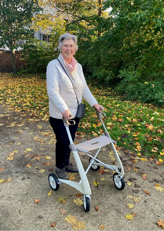 Smiling elderly woman standing outdoors with a modern four-wheeled walker on a leafy path in autumn.