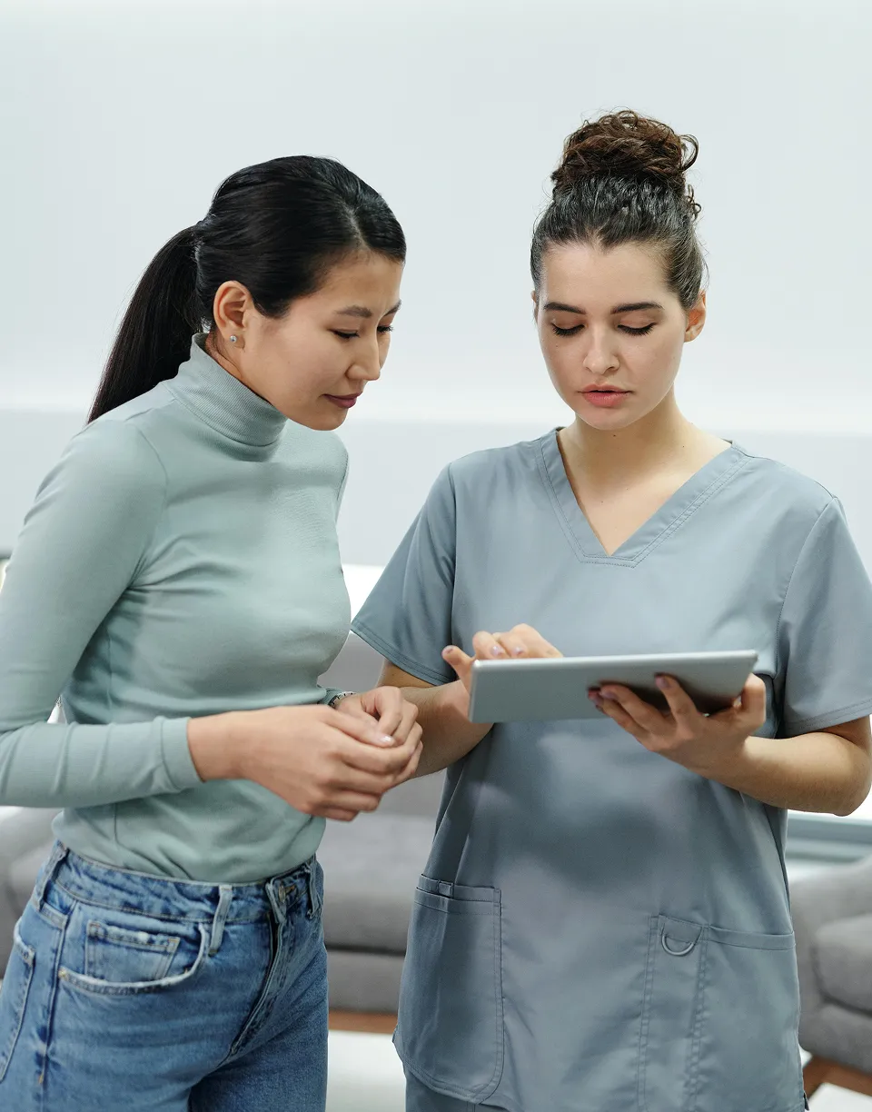 A nurse in scrubs shows information on a tablet to a woman in a green turtleneck and jeans.