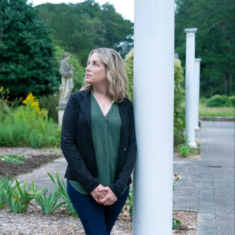 A woman with blonde hair stands outdoors beside a white column, looking to her left, with a garden and statue in the background.
