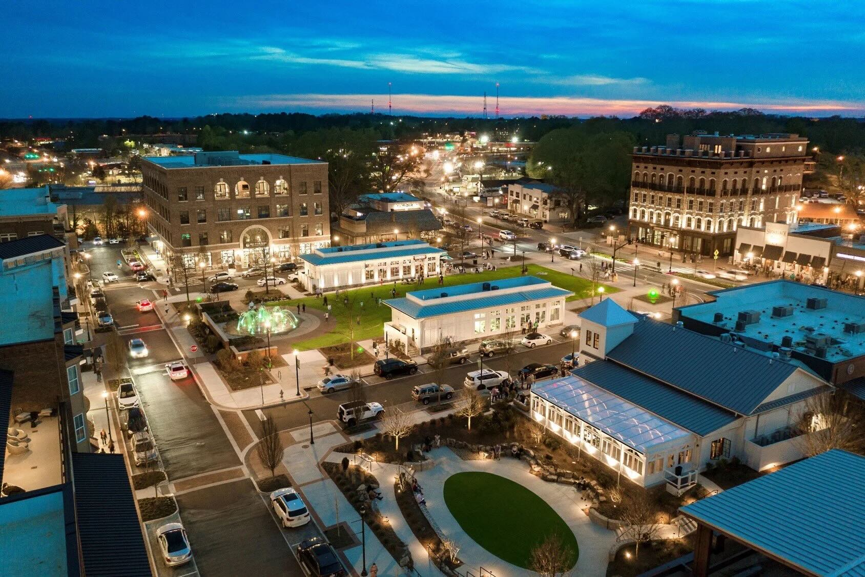 Evening view of downtown Alpharetta, Georgia, with lit-up buildings, a central green space, and busy streets filled with cars and people.