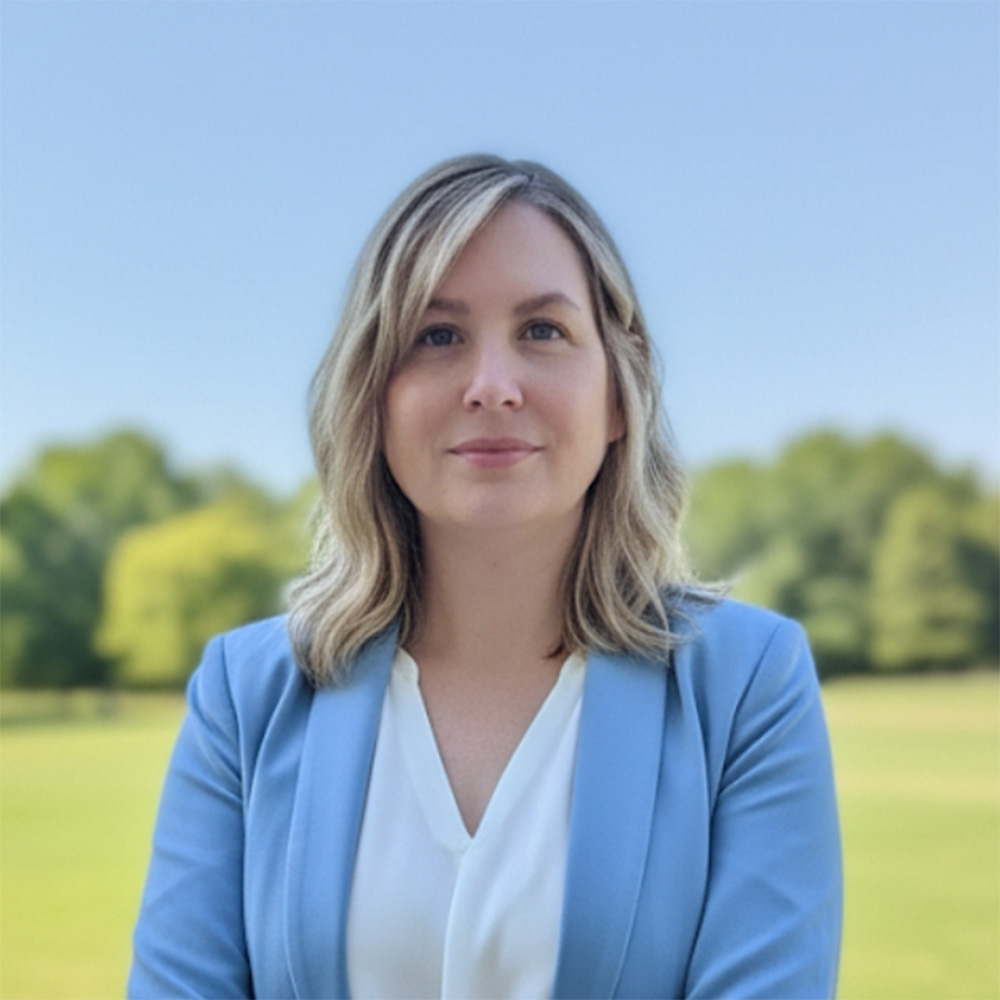Portrait of a woman with shoulder-length blonde hair in a light blue blazer and white blouse, standing outdoors in a sunny park with trees and a clear blue sky.