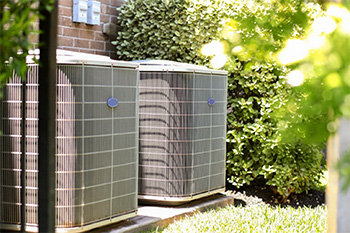 Two large outdoor central air conditioning units installed side by side on concrete pads next to a brick wall, surrounded by green shrubs and sunlight.