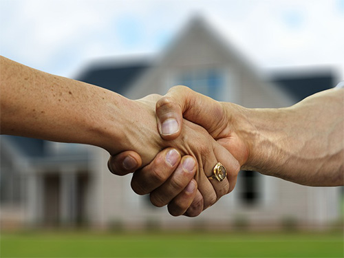 Two people shake hands in front of a house, symbolizing a real estate deal or agreement.