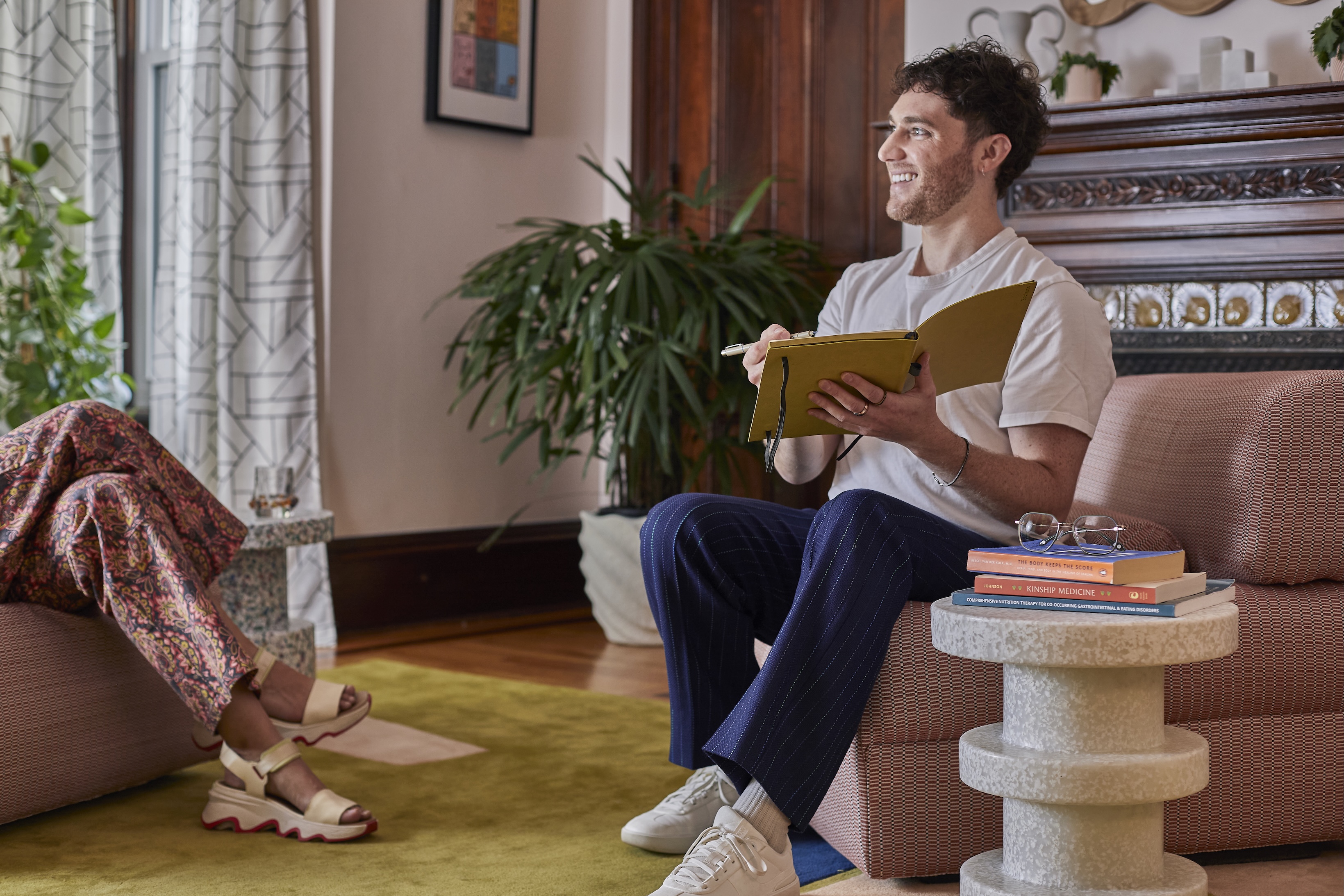 Man smiling and writing in a yellow notebook while sitting on a patterned couch during a conversation in a cozy room.