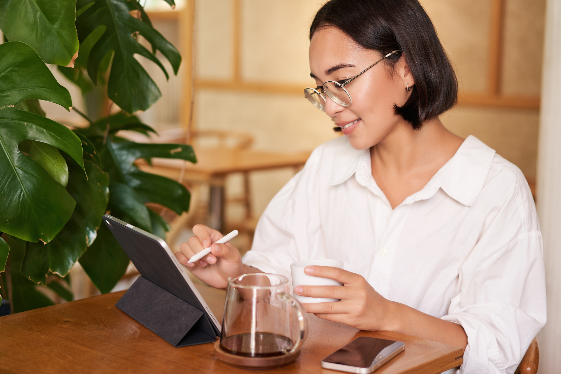 Person seated at a wooden table in a café, using a tablet with a stylus while holding a cup of coffee, with a smartphone and glass carafe nearby and greenery in the background.