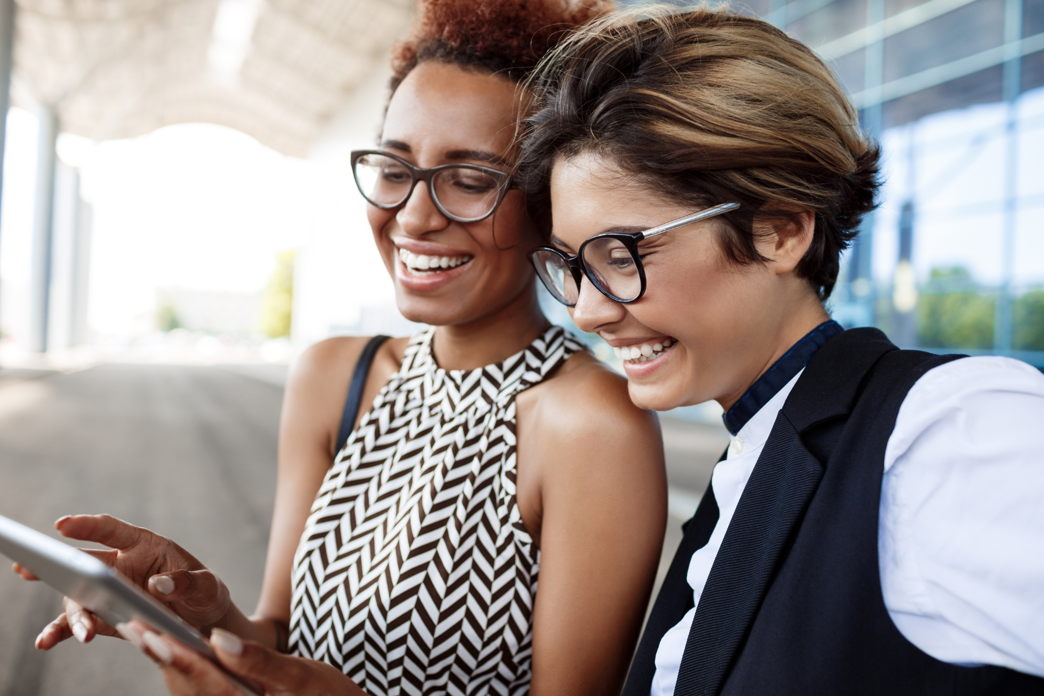 Two women smiling while reviewing content on a tablet together outdoors
