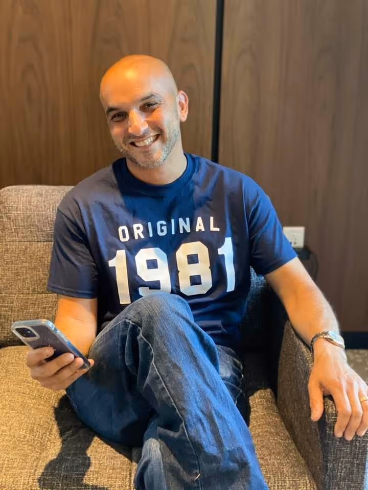 Kris Torres smiling while sitting on a couch and holding a phone, wearing a navy “Original 1981” shirt