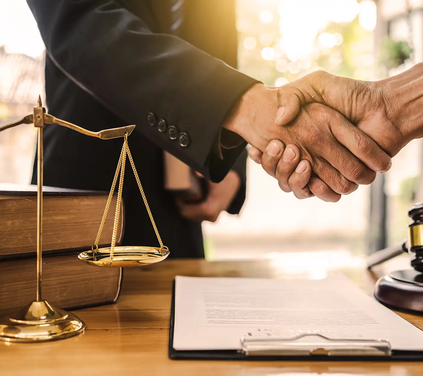 Close-up of a handshake between two people in a legal office with scales of justice, law books, and a contract on a desk.