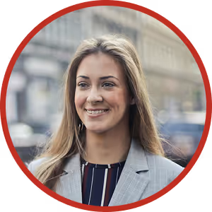 Smiling young woman with long blonde hair wearing a gray blazer and striped shirt in an urban setting.
