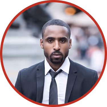 Serious man with short hair and beard wearing a black suit and tie, outdoors in an urban setting.