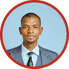 Portrait of a serious man wearing a gray suit, white shirt, and patterned tie against a light blue background with a red circular border.