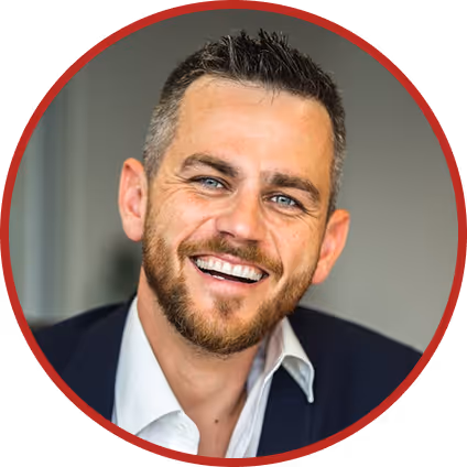 Smiling man with short dark hair and beard wearing a white shirt and dark blazer against a blurred background.