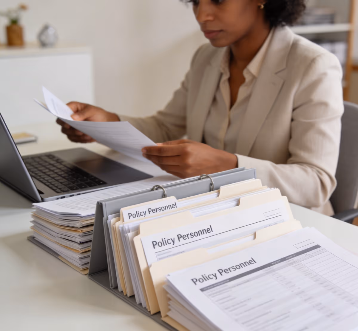 Woman reviewing documents at desk with open laptop and organized file folders labeled Policy Personnel.