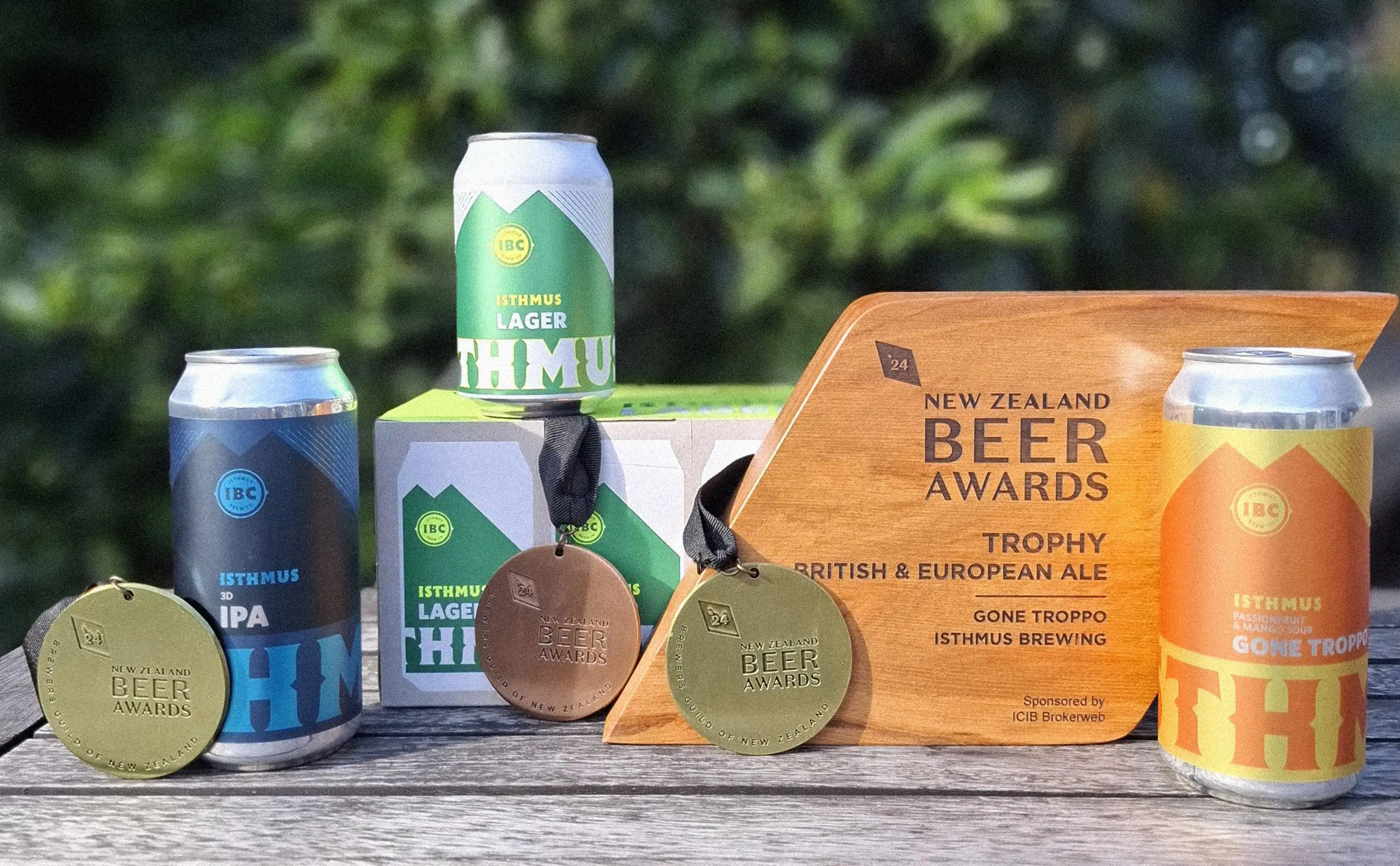 Four cans of Isthmus beer, three medals, and a wooden New Zealand Beer Awards trophy are displayed on a table outdoors, with green foliage blurred in the background.
