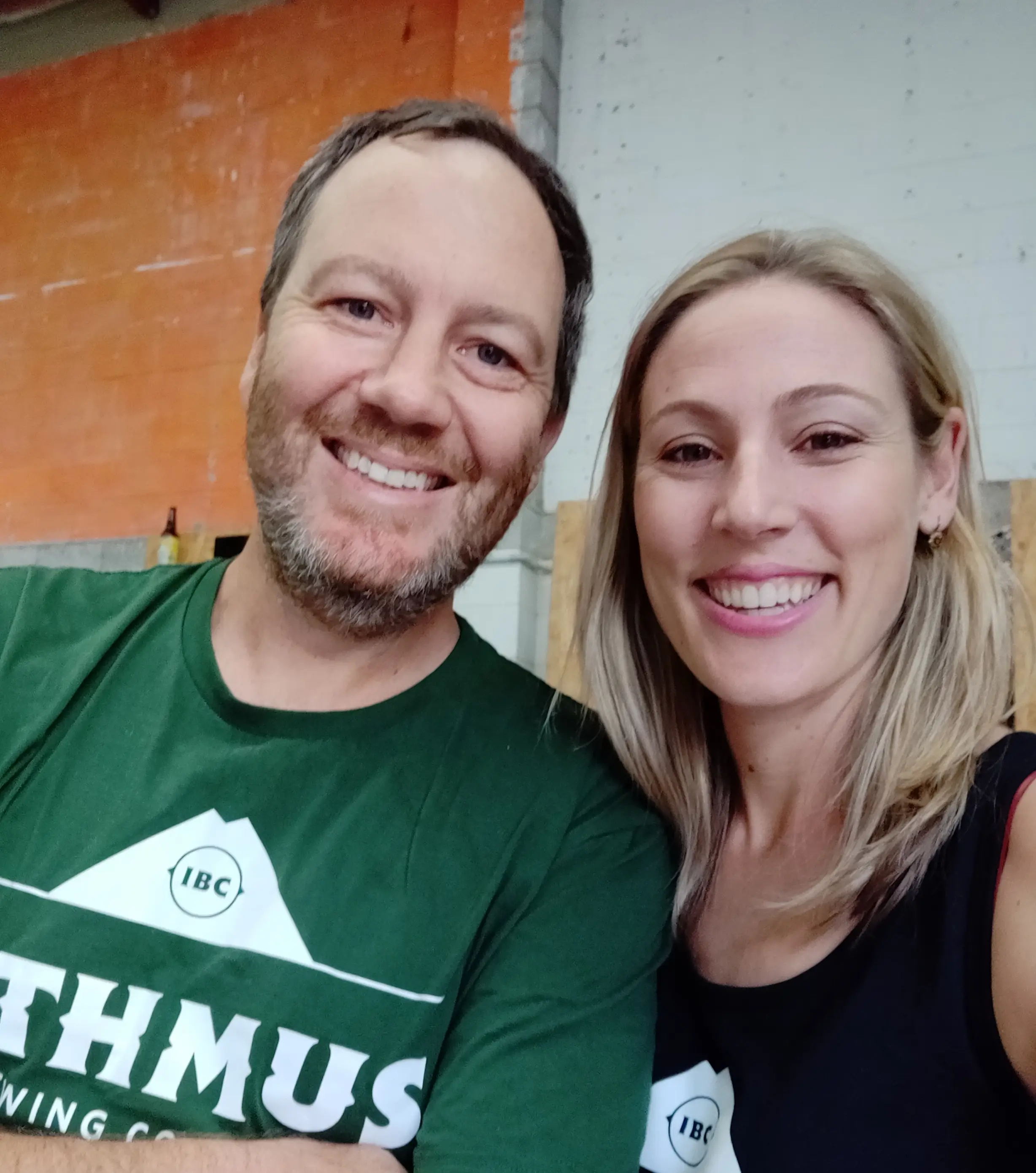 Two smiling adults take a selfie indoors. The man has short brown hair and a beard, wearing a green t-shirt, while the woman has long blonde hair and is wearing a black tank top. The background shows an orange and white wall.
