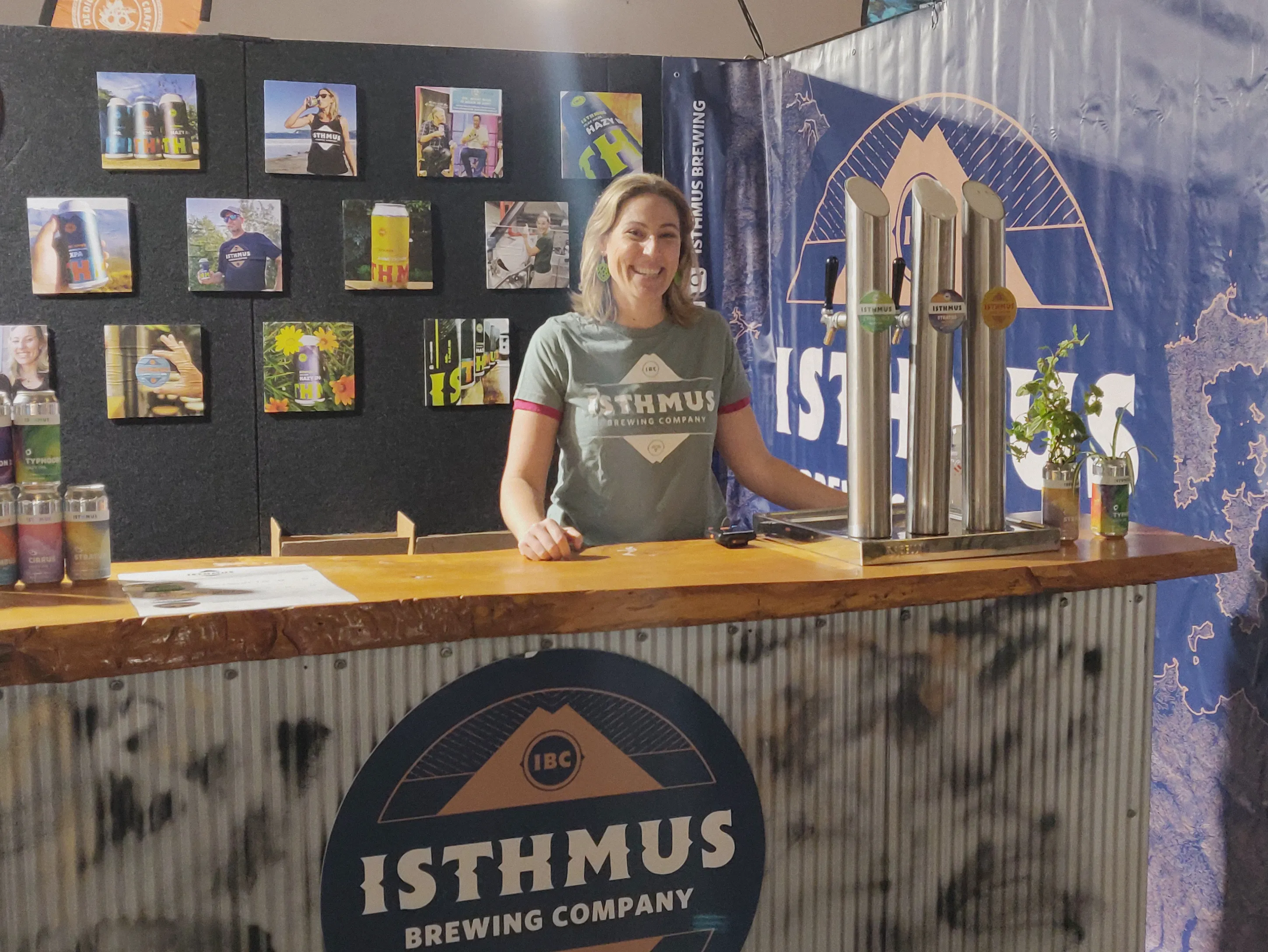 A smiling woman wearing an Isthmus Brewing Company t-shirt stands behind a wooden bar with beer taps. The bar displays the Isthmus logo, and colorful cans and photos decorate the booth in the background.