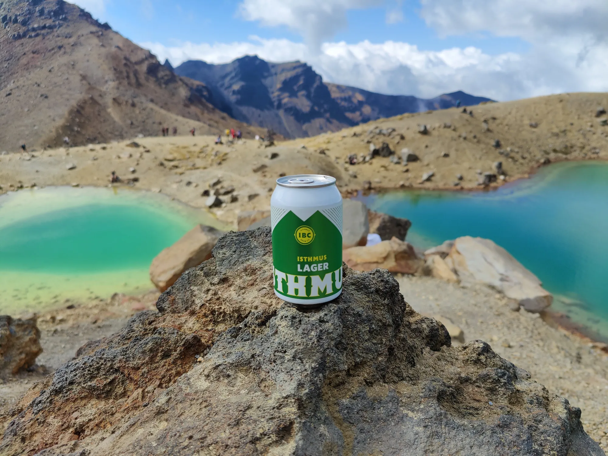 A can of Isthmus Lager beer sits on a rocky surface in front of turquoise lakes and rugged, mountainous terrain under a partly cloudy sky.