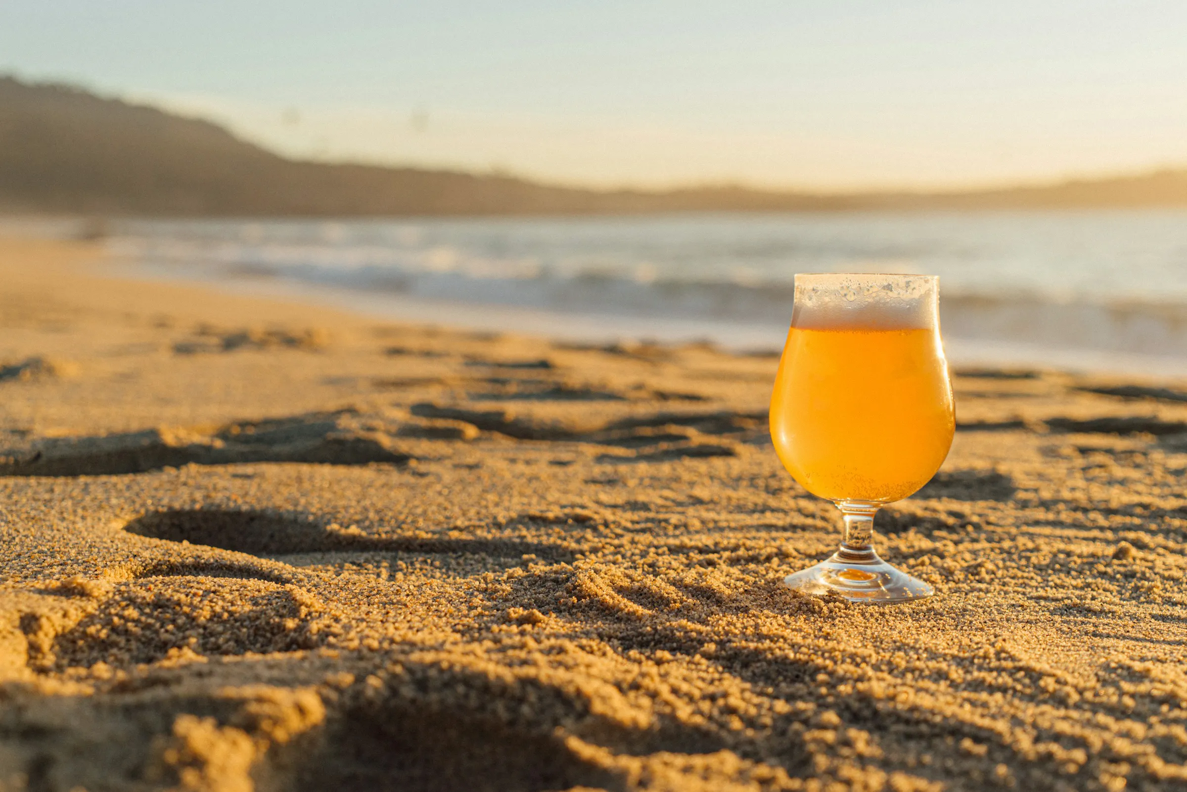 A glass of beer sits on sandy beach at sunset, with gentle waves and distant hills in the background under a golden sky.