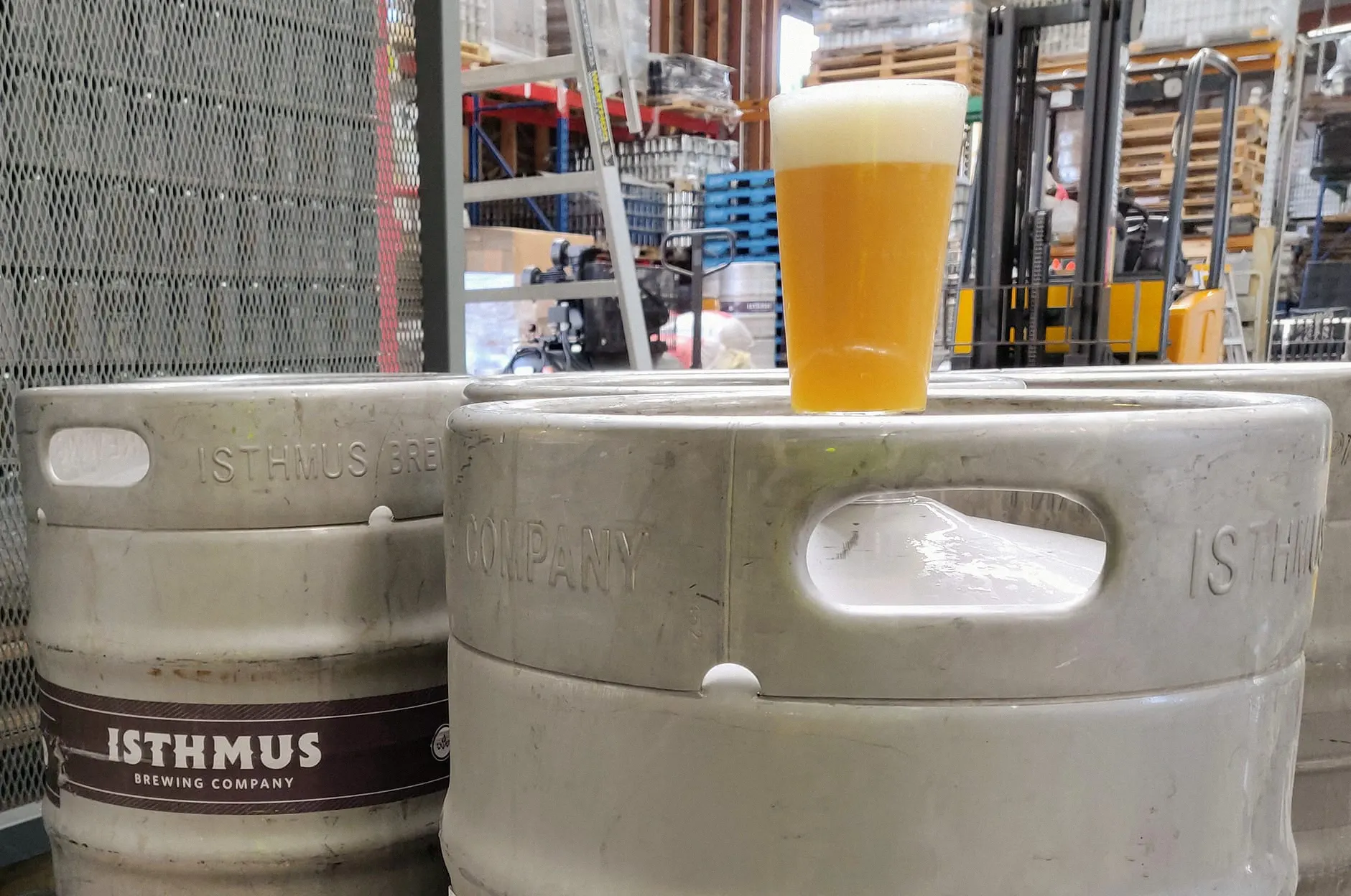 A pint of hazy beer with a thick foamy head sits on top of a metal keg in a brewery warehouse, surrounded by stacked kegs and pallets.