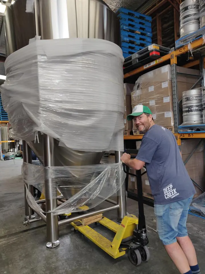 A man in a cap and Deep Creek t-shirt uses a yellow pallet jack to move a large, plastic-wrapped stainless steel tank in a warehouse with shelves and beer kegs.