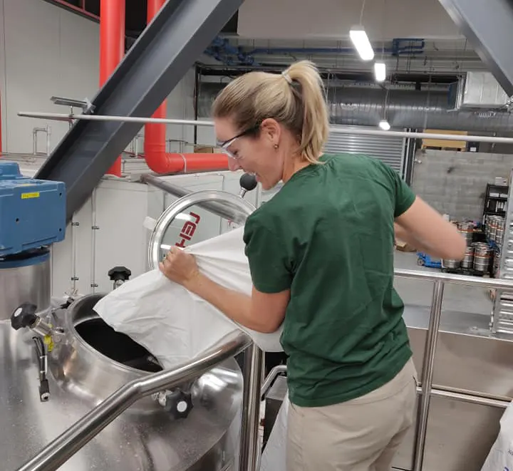 A woman wearing glasses and a green shirt pours ingredients from a large white bag into a metal brewing tank inside a modern brewery.