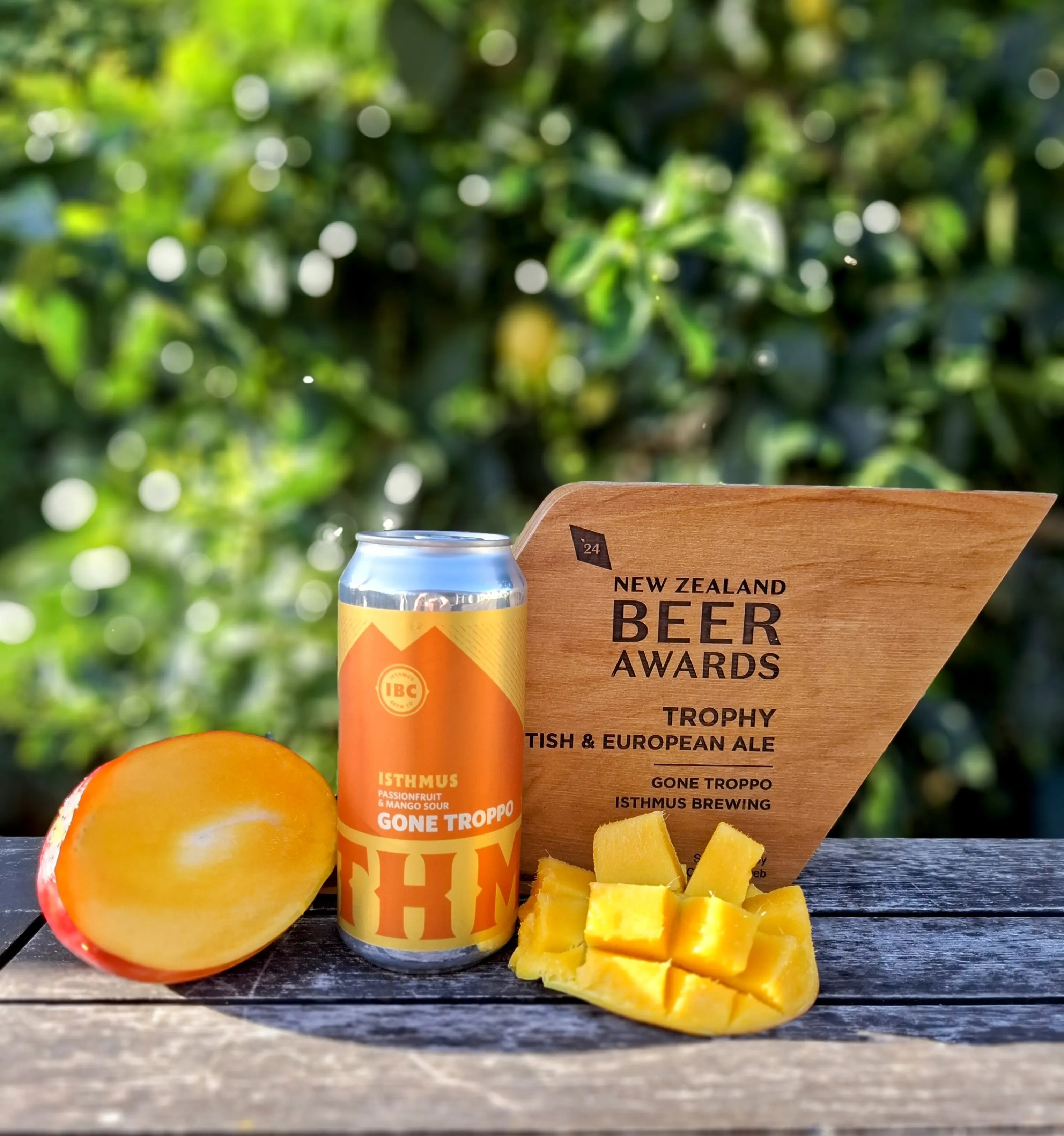 A can of Isthmus Gone Troppo ale, a sliced mango, and a New Zealand Beer Awards trophy sit on a wooden surface outdoors, with green foliage blurred in the background.