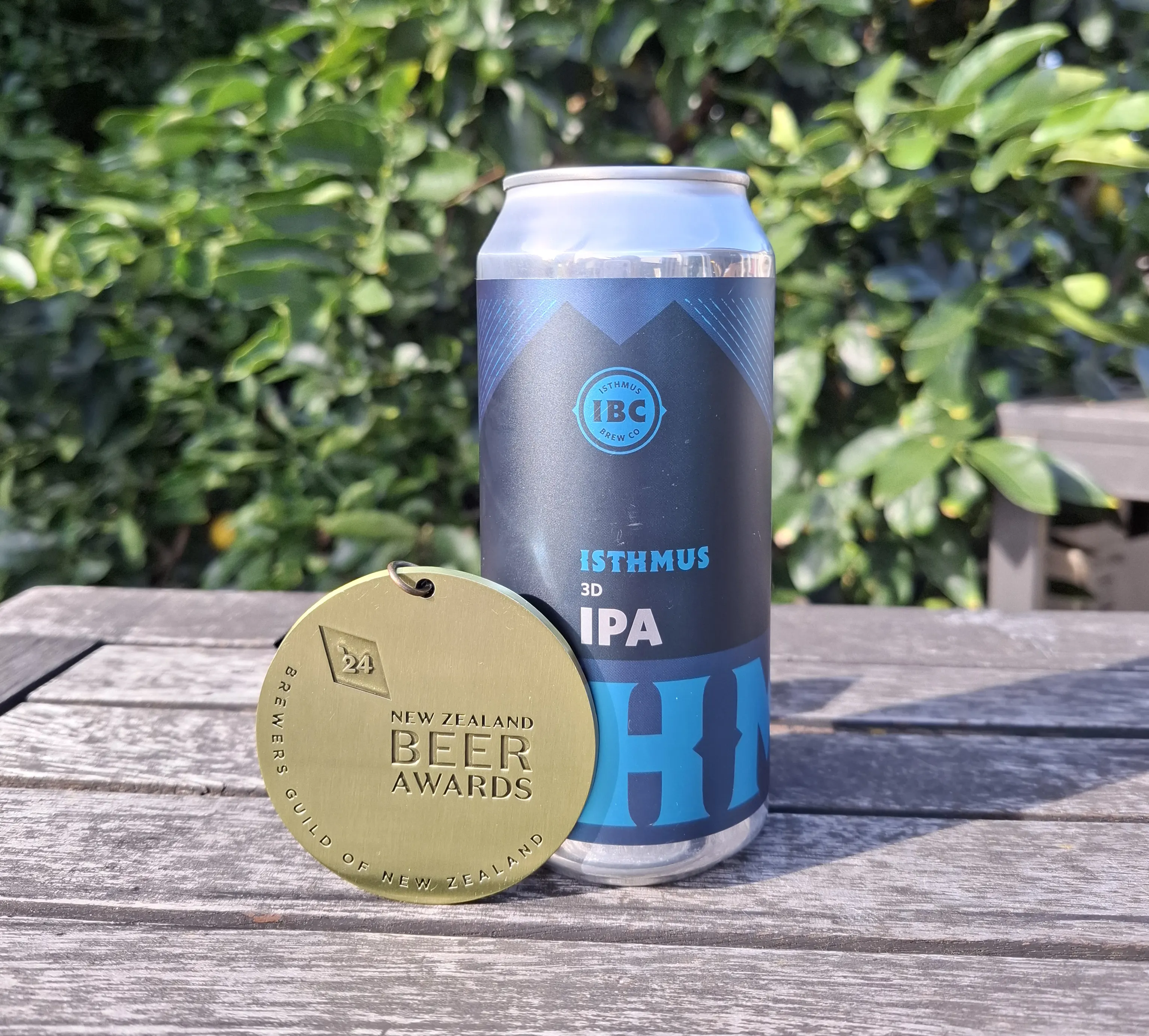 A can of Isthmus 3D IPA beer sits on a wooden table next to a gold New Zealand Beer Awards medal. Lush green foliage is visible in the background.