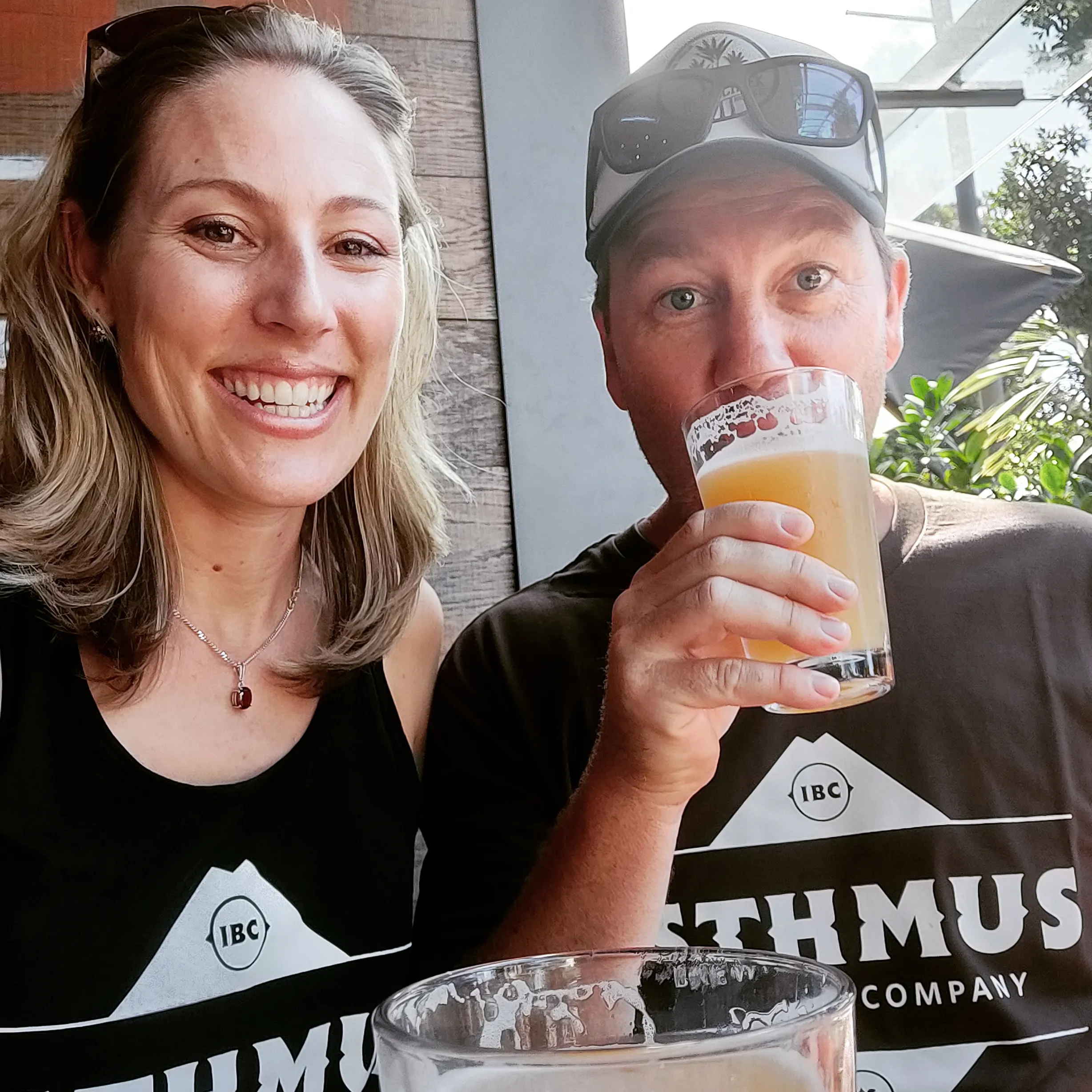 A smiling woman and a man wearing a cap sit together. The man is drinking a beer. Both are wearing matching black Isthmus Beer Company shirts. They appear to be at an outdoor or semi-outdoor setting.