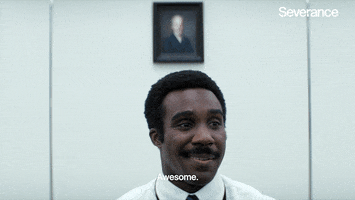 Man in white shirt and tie with a mustache smiling and saying 'Awesome' inside an office with a blurred portrait on the wall behind him.