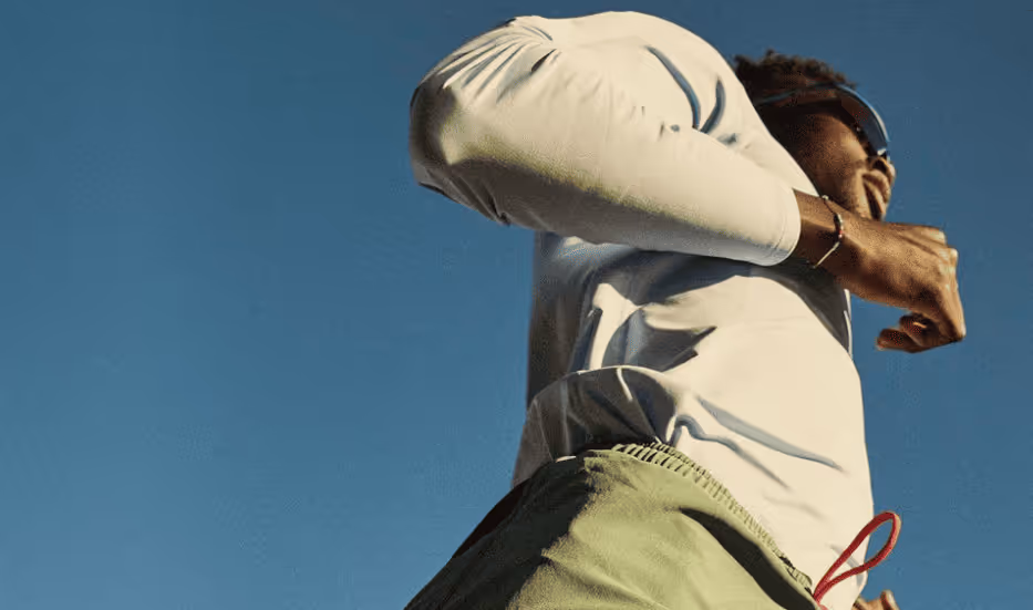 Man jogging outdoors wearing sunglasses and a light-colored long-sleeve shirt under a clear blue sky.
