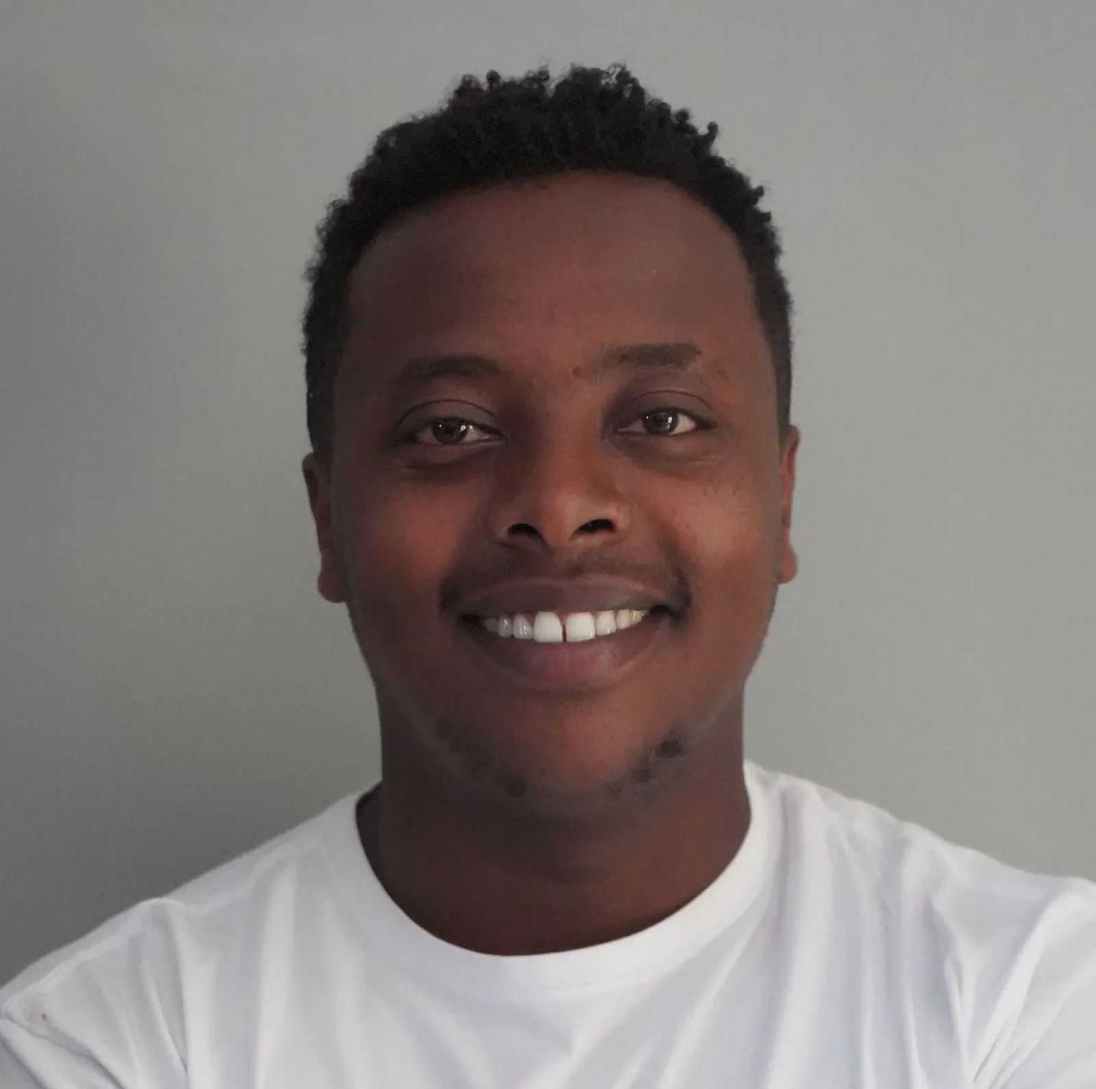 Smiling man with short curly hair wearing a white shirt standing against a gray background.