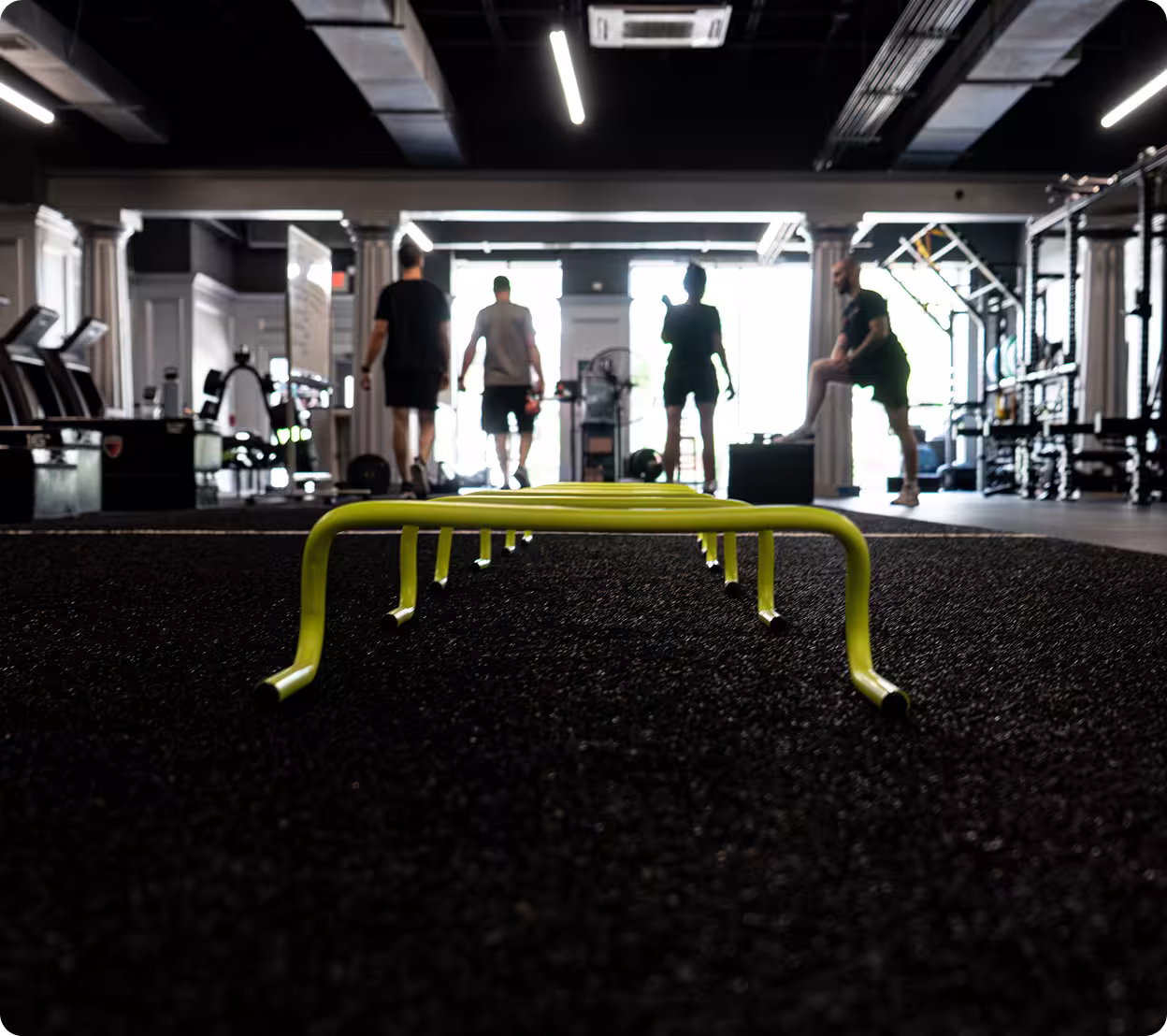 a yellow agility hurdle on a dark, textured gym floor, with four people exercising in the background