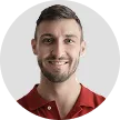 Smiling young man with short dark hair and beard wearing a red shirt against a light gray background.