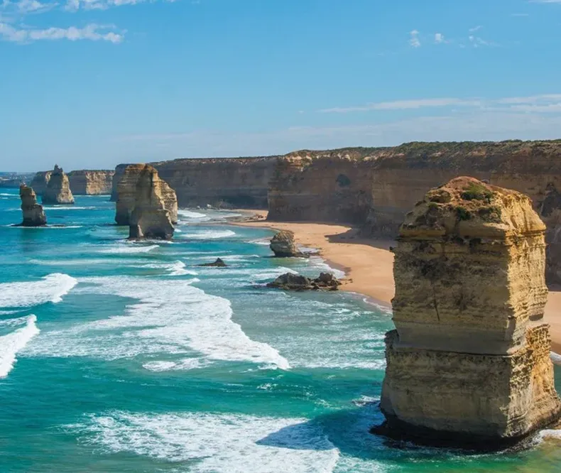 The Twelve Apostles limestone rock formations rising from the Southern Ocean along the coastline of a sandy beach under a blue sky.