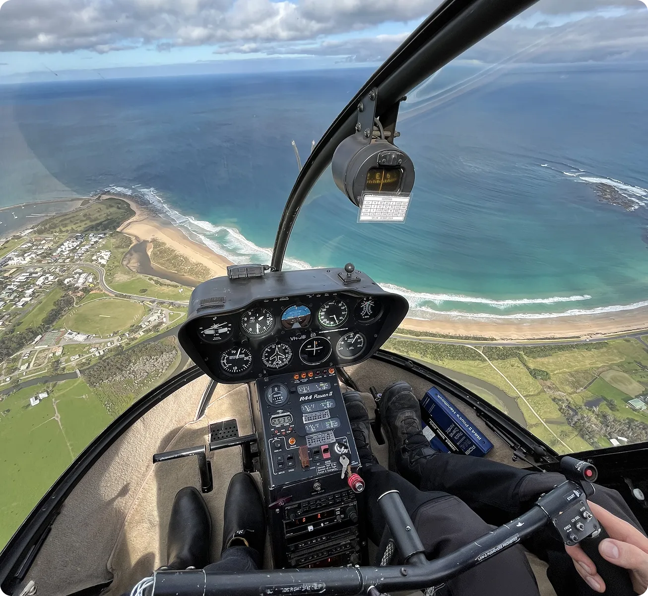 View from inside a helicopter cockpit flying over a coastline with clear blue water and sandy beaches.