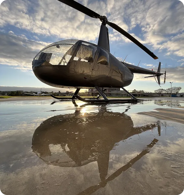Black helicopter parked on a wet concrete surface, reflecting in a puddle with a partly cloudy sky background.