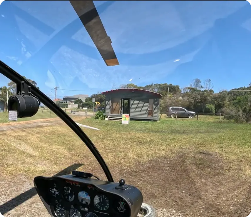 View from inside a helicopter cockpit looking out at a small house and a parked SUV on grassy land under a clear blue sky.