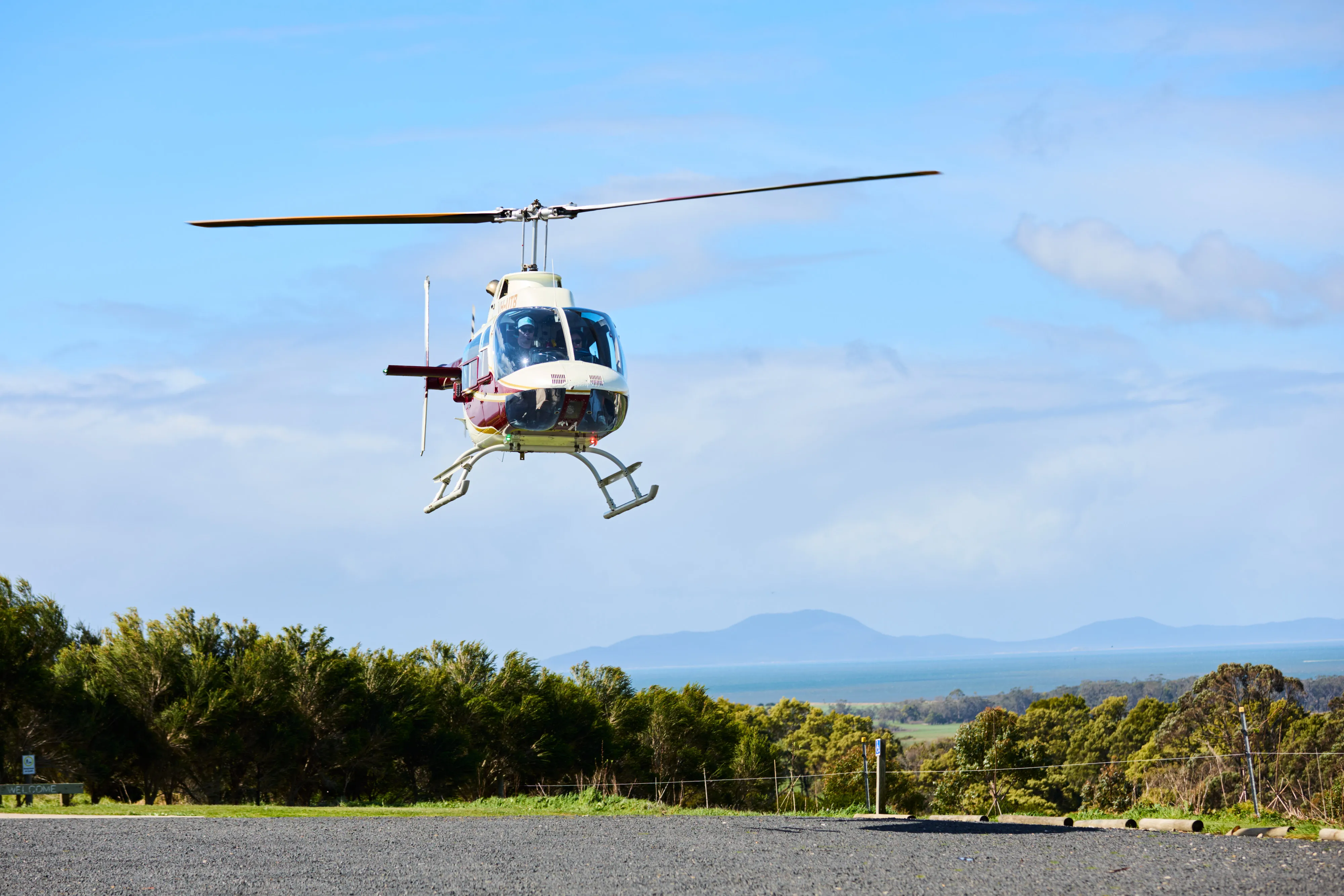 White and maroon helicopter hovering above a gravel surface with trees and distant mountains under a blue sky.