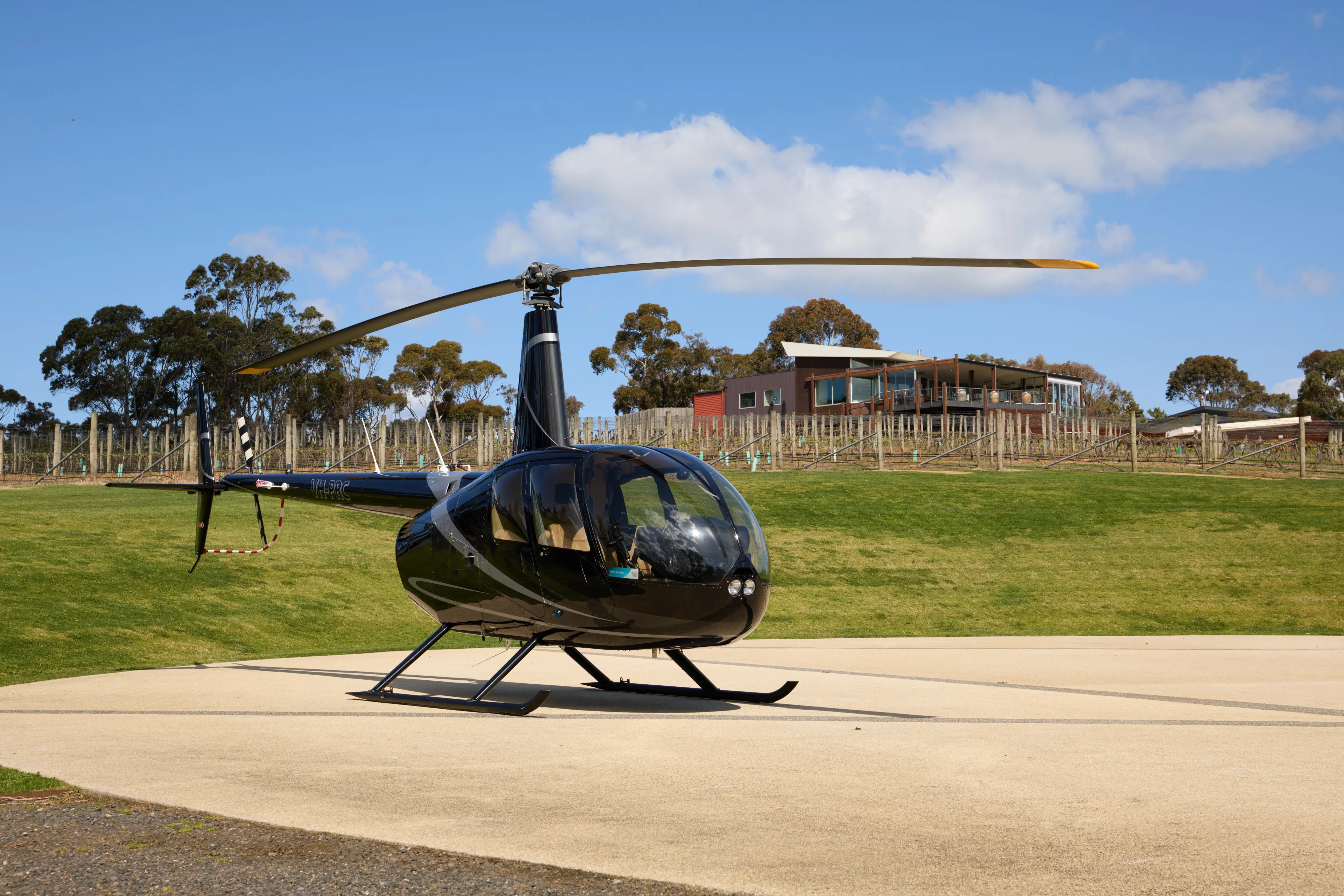 Black helicopter parked on a concrete helipad with green grass, a vineyard, and a modern building in the background under a blue sky.