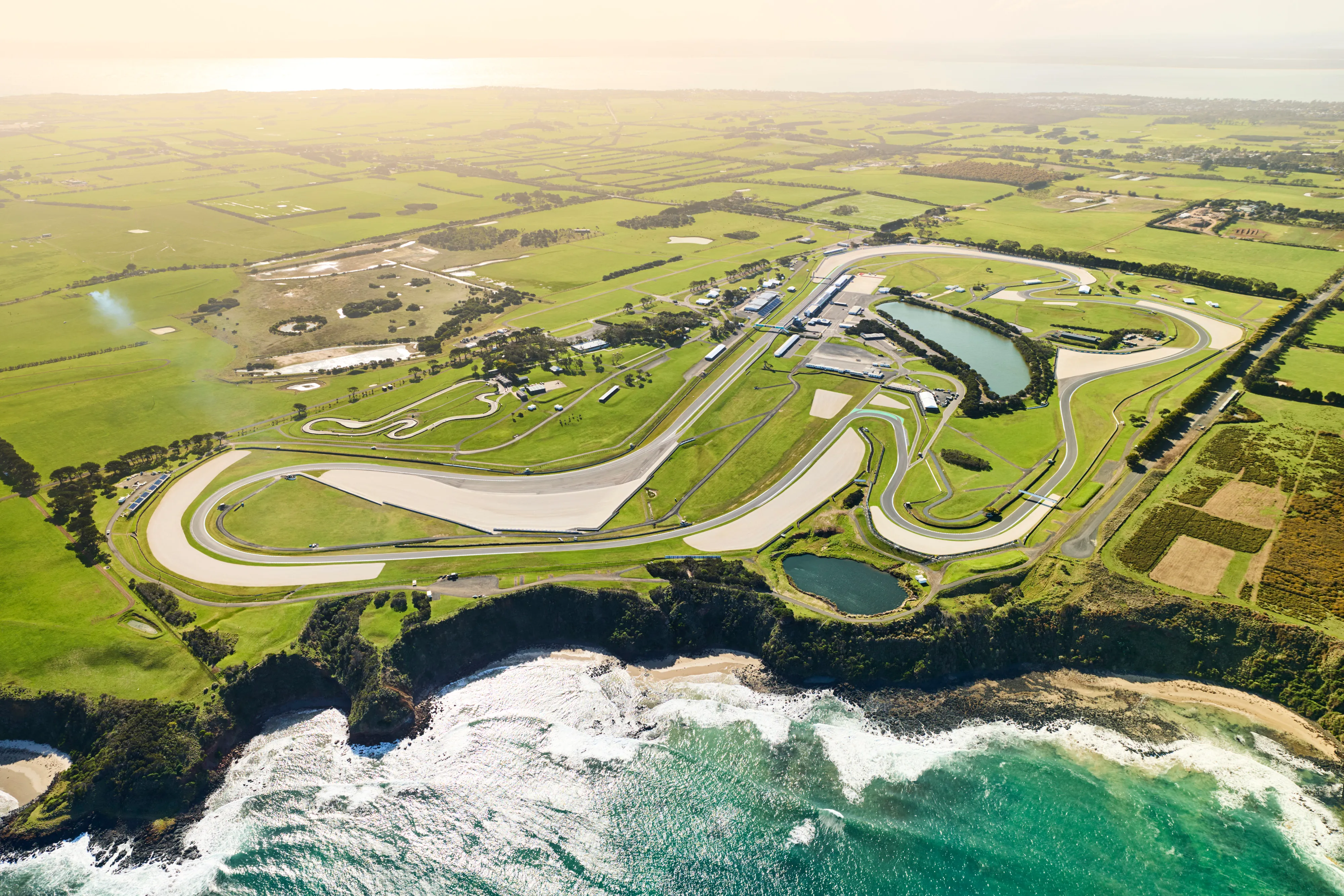 Aerial view of a coastal racing circuit with winding tracks surrounded by green fields and cliffs overlooking the ocean.