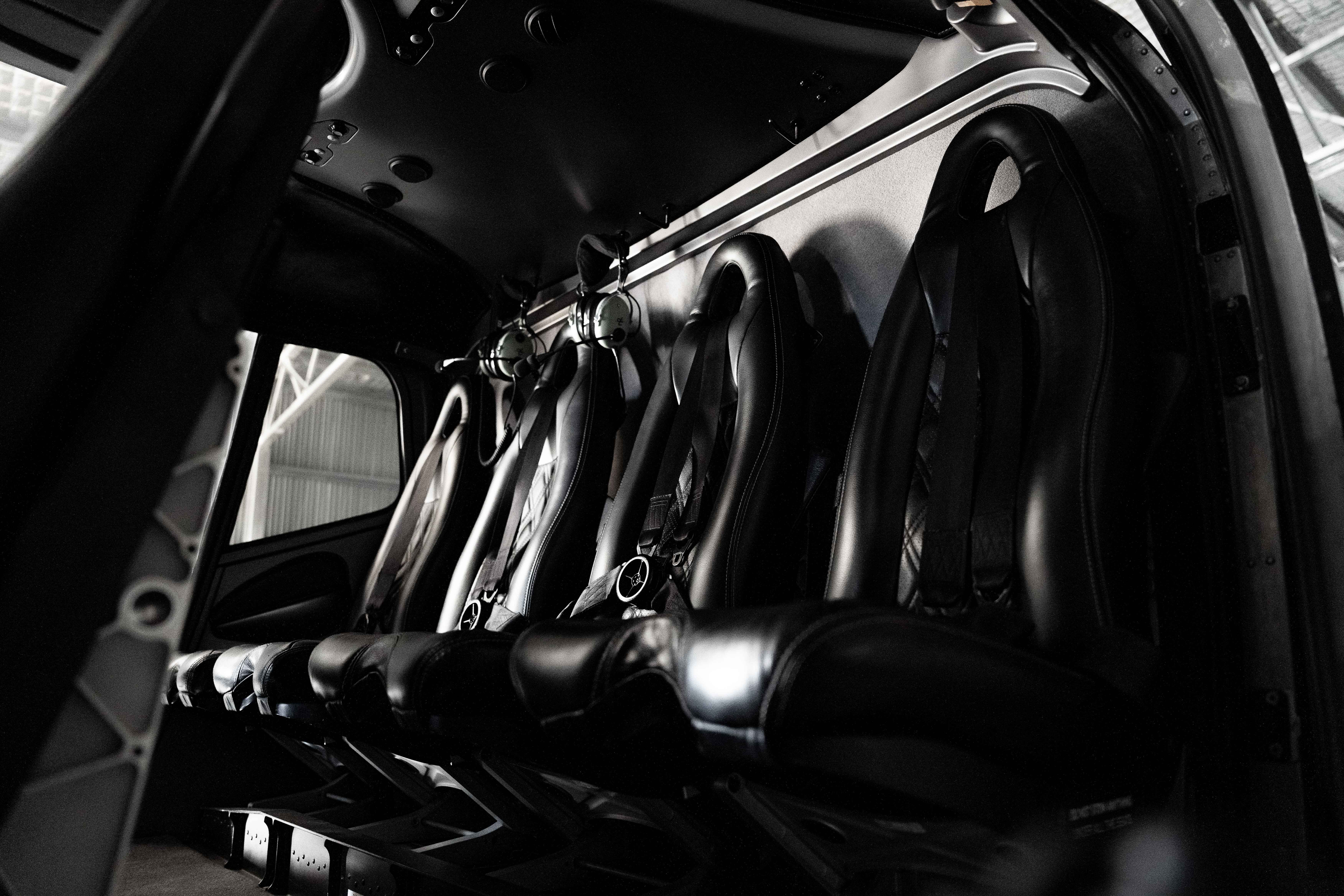 Interior view of a helicopter cabin showing a row of five black leather seats with seatbelts and hanging headsets.