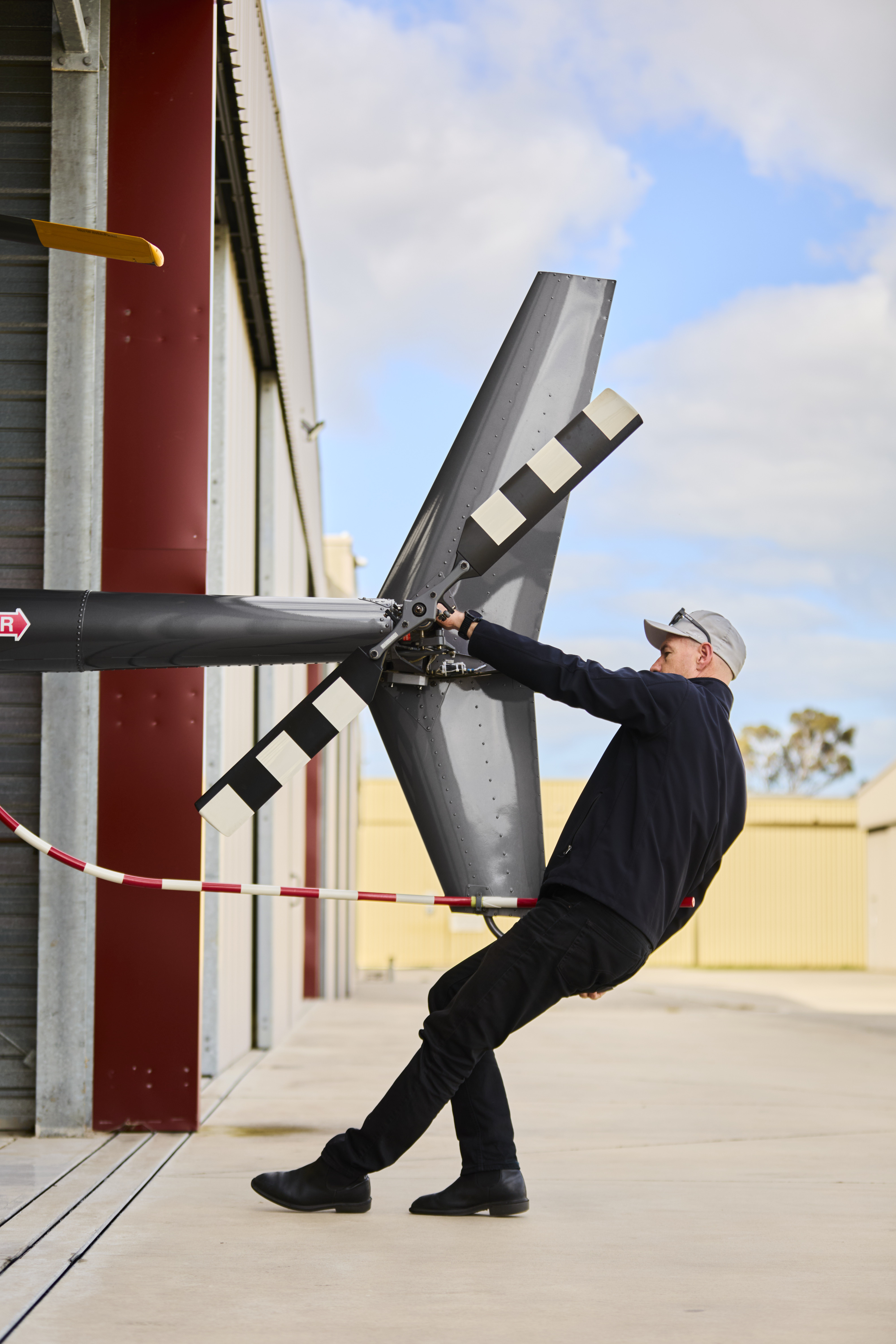 Man wearing black clothing and gray cap leaning back while holding a helicopter's tail rotor blades outside near a hangar.