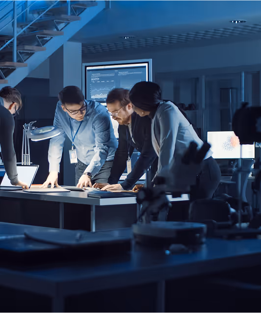Group of professionals in an office at night collaborating over documents on a table with computer screens in the background.