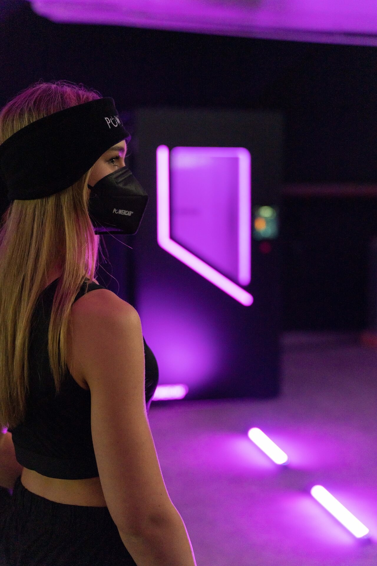 Frau mit schwarzer Powercap-Stirnband und schwarzer Maske in einem Raum mit violetter Beleuchtung.