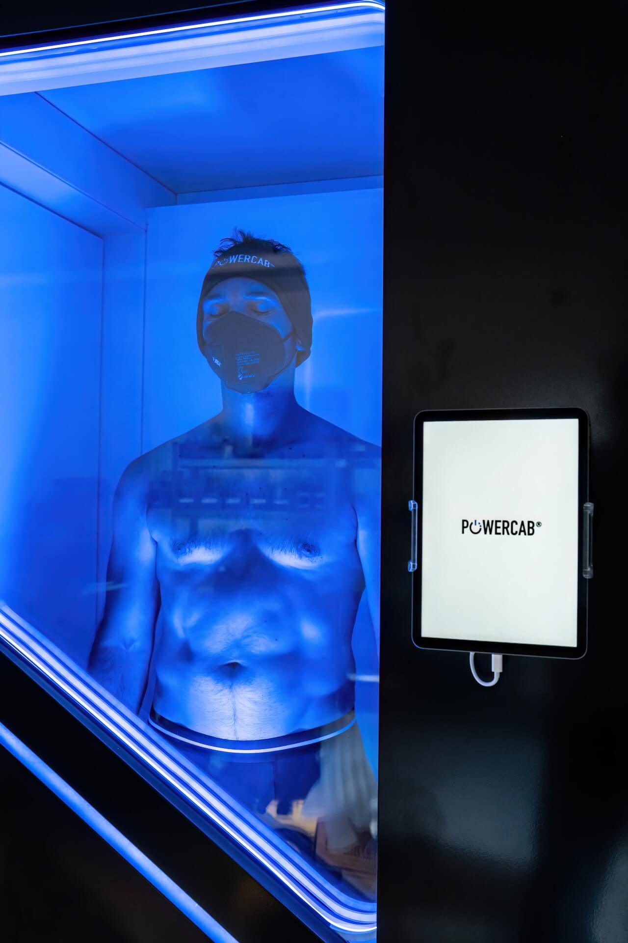 Mann mit geschlossenen Augen trägt eine Maske und Mütze und steht in einer blau beleuchteten Kabine neben einem Bildschirm mit der Aufschrift POWER CAB.