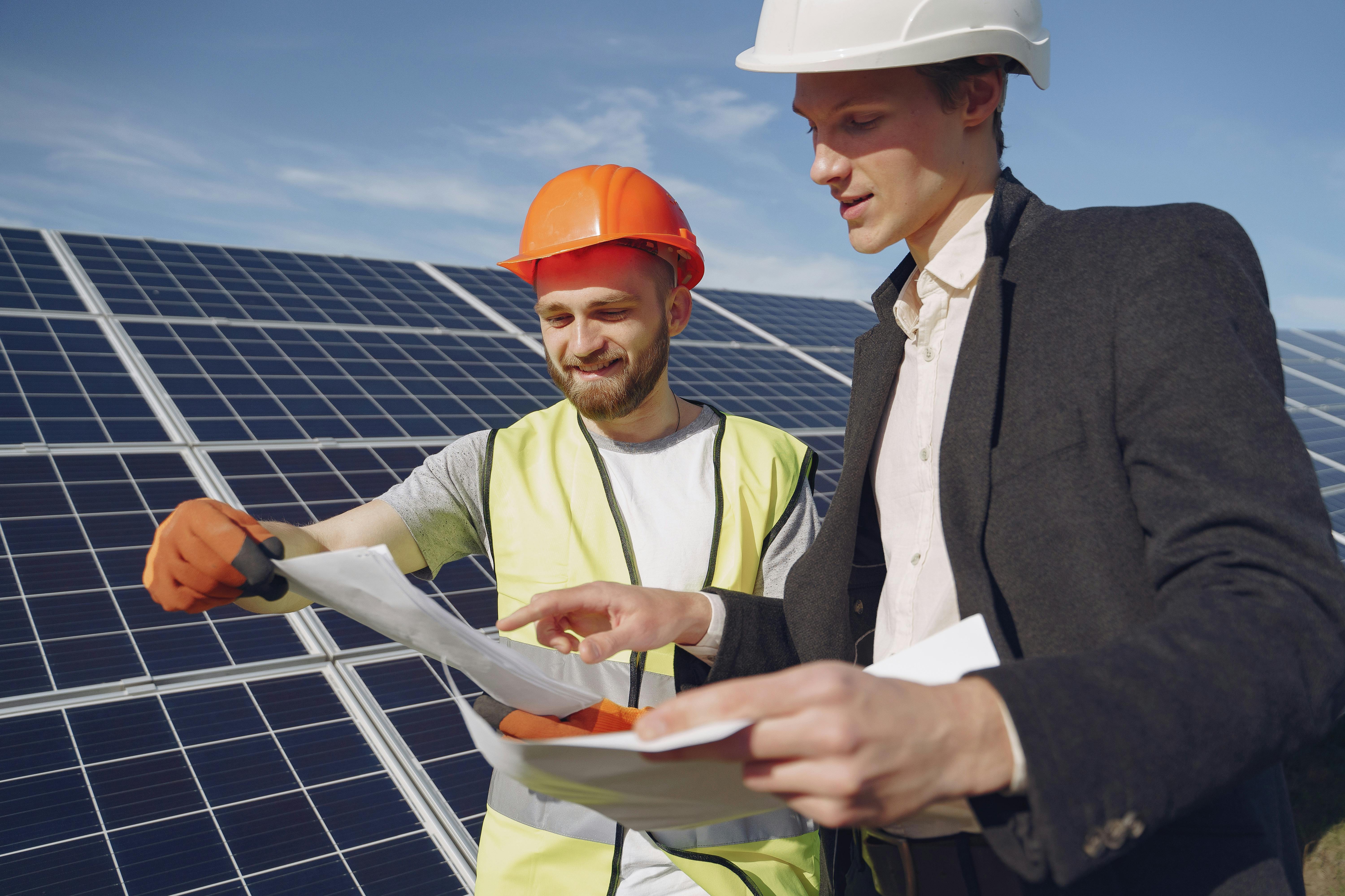 Two men are standing outside on a sunny day with a few clouds in the sky. They're standing in front of an array of solar panels. They're either looking at a blueprint or order forms. One man is quite tall wearing a suit, and the other man is wearing an orange hard hat and a yellow PPE vest.
